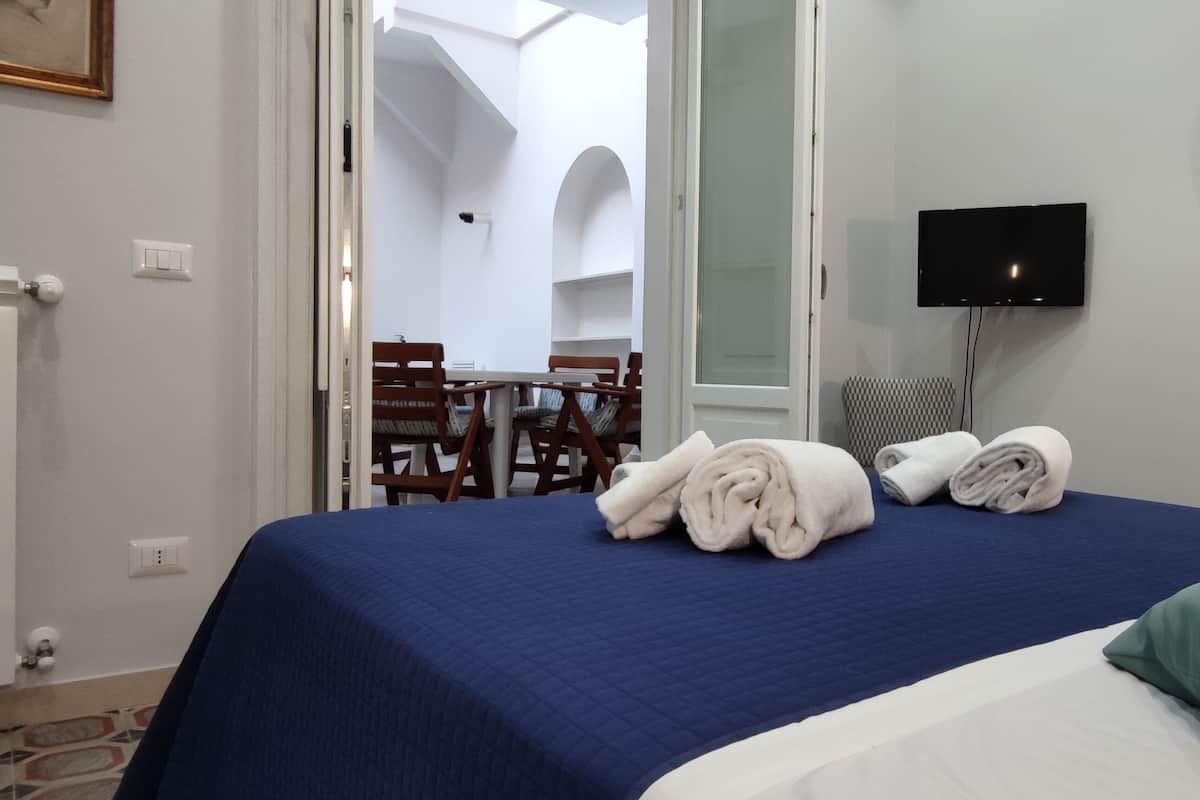A hotel room with a bed, a TV, a mirror, and a view of an outdoor patio with chairs and a table.