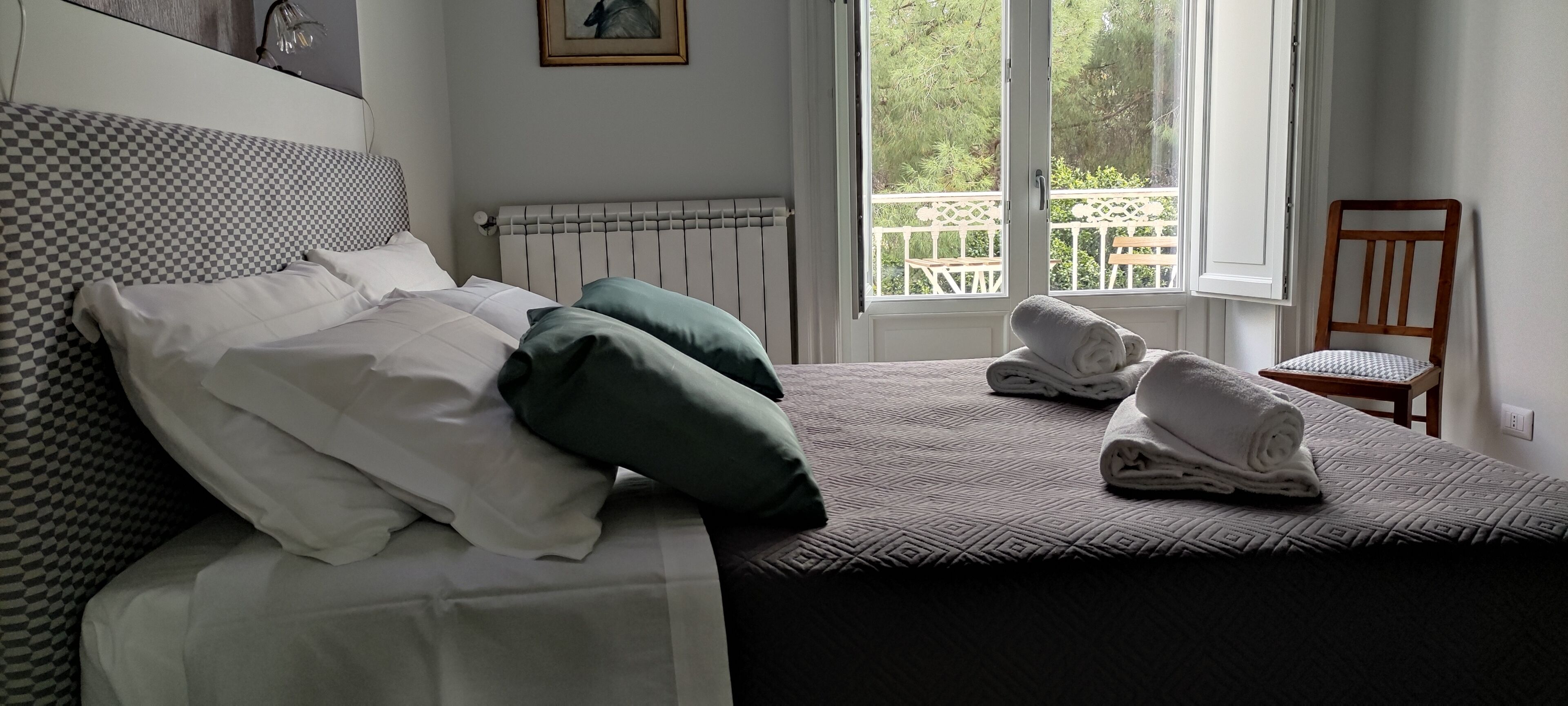 Una cama bien hecha con almohadas, una toalla doblada y una silla junto a la ventana.