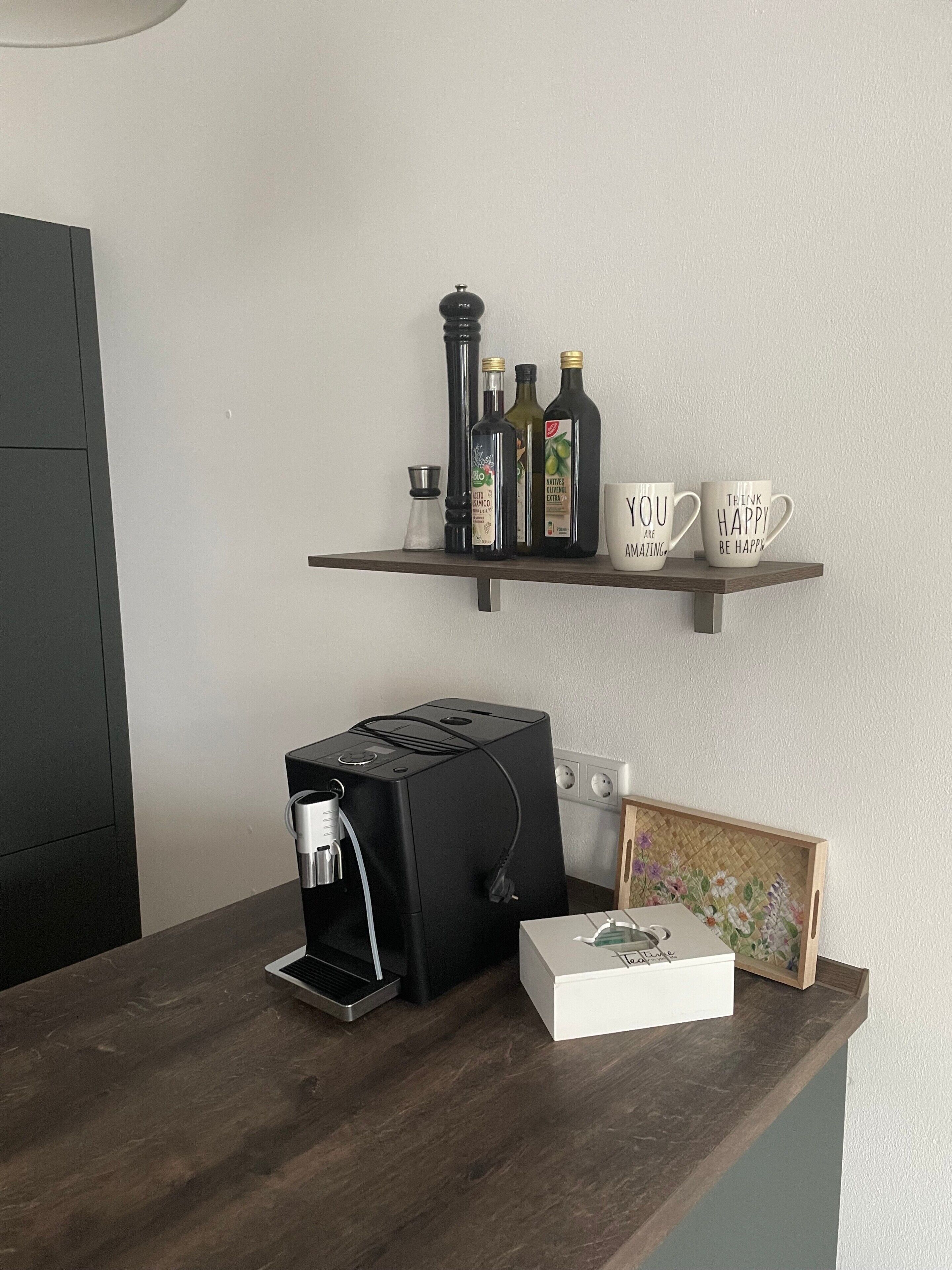 A coffee maker, a box, and a framed picture on a wooden table with a shelf above containing bottles and mugs.