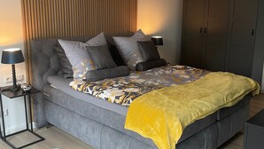 Un dormitorio moderno con un sofá cama gris, una pared de listones de madera, dos lámparas colgantes blancas, una manta amarilla y una alfombra blanca con forma de nube.