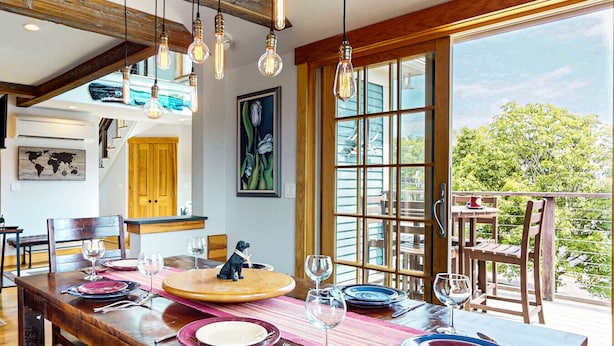 A dining area with a large wooden table set for a meal, a dog figurine centerpiece, and a view of greenery through large windows.