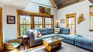 A modern living room with a sectional sofa, a glass coffee table, and large windows offering a view of the outdoors.