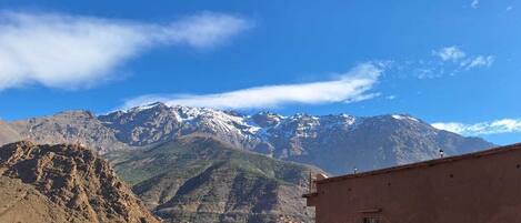 A mountainous landscape with snow-capped peaks in the background, a building with a flat roof in the foreground, and a clear blue sky.