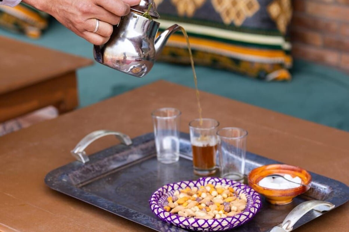 A tray with a teapot pouring tea, glasses, and a bowl of nuts on a table.
