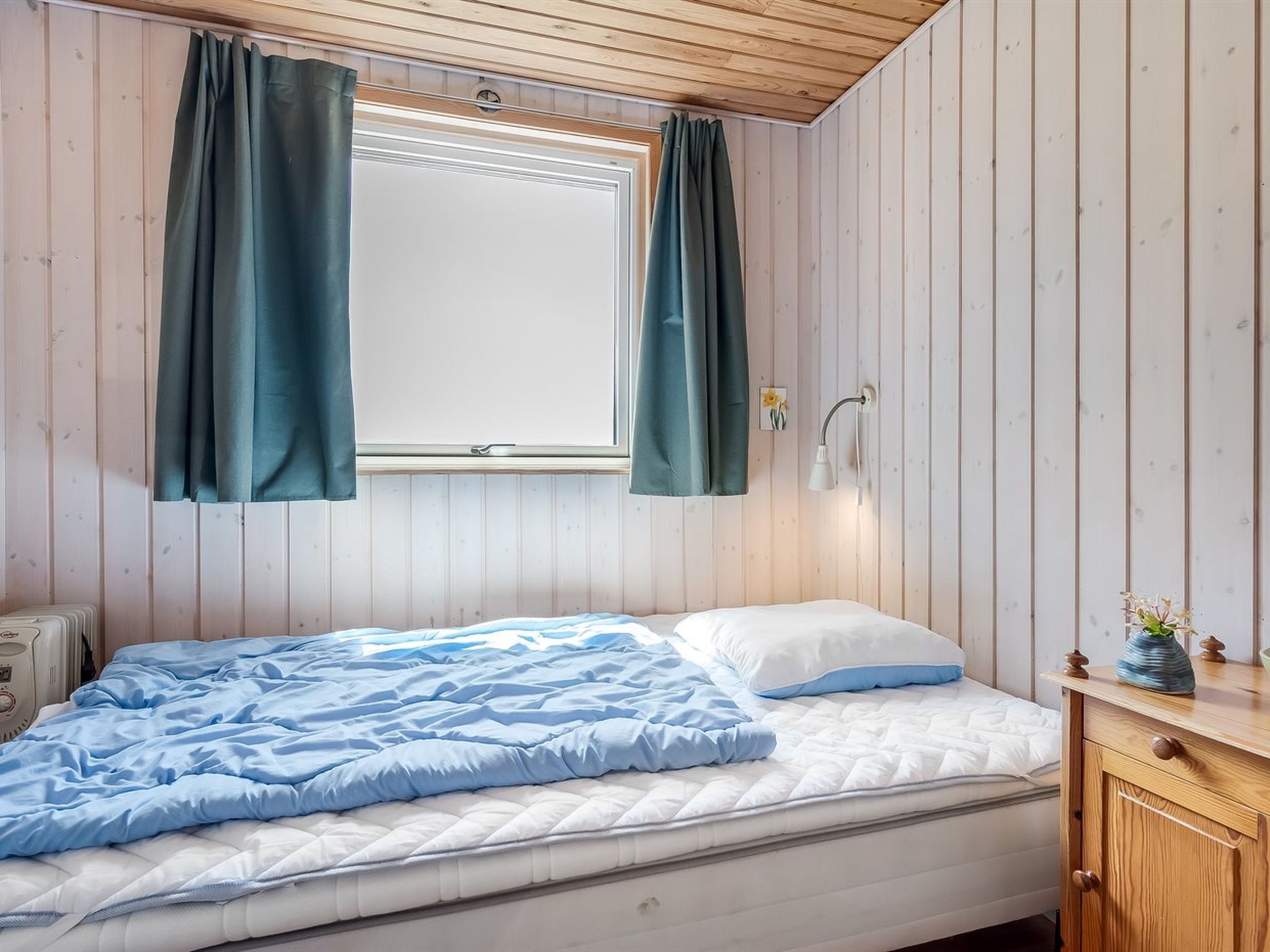 Un dormitorio con cama, ventana con cortinas, lámpara de noche y cómoda de madera.