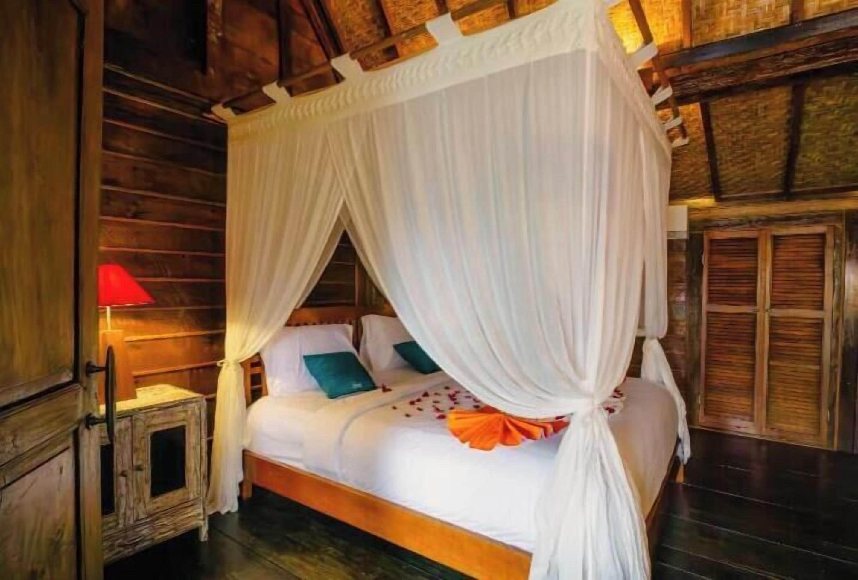 Ein Zimmer mit Holzboden, einem Himmelbett, einem Nachttisch mit Lampe und einer Holztür.