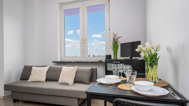 A modern living room with a grey sofa, a dining table with plates and glasses, a TV on a stand, and a window with a city view. - Apartamenty Sun & Snow Wałowa (Warsaw)