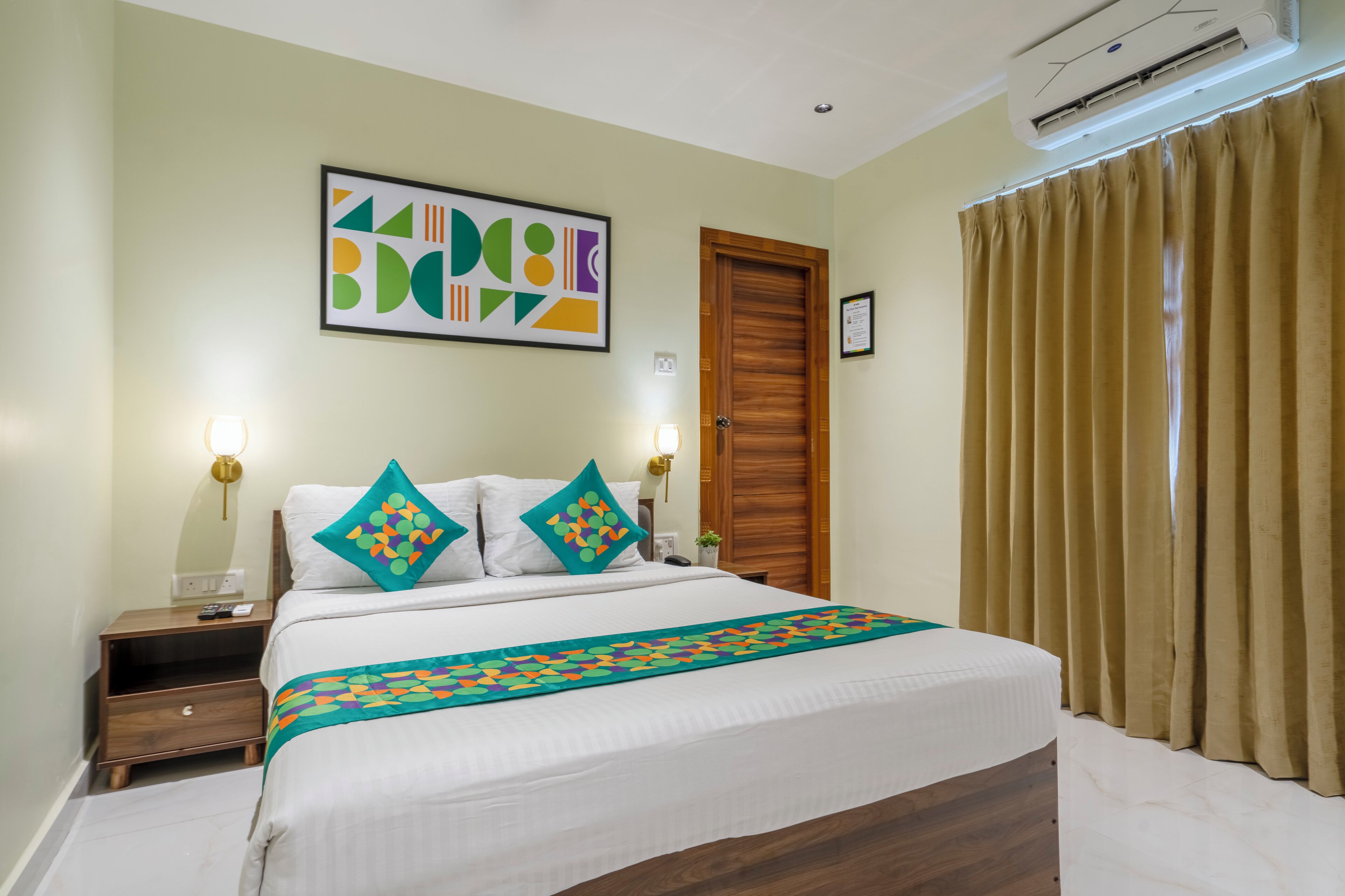 A hotel room with a bed, a nightstand, a wall-mounted air conditioner, and a door with a wooden frame.