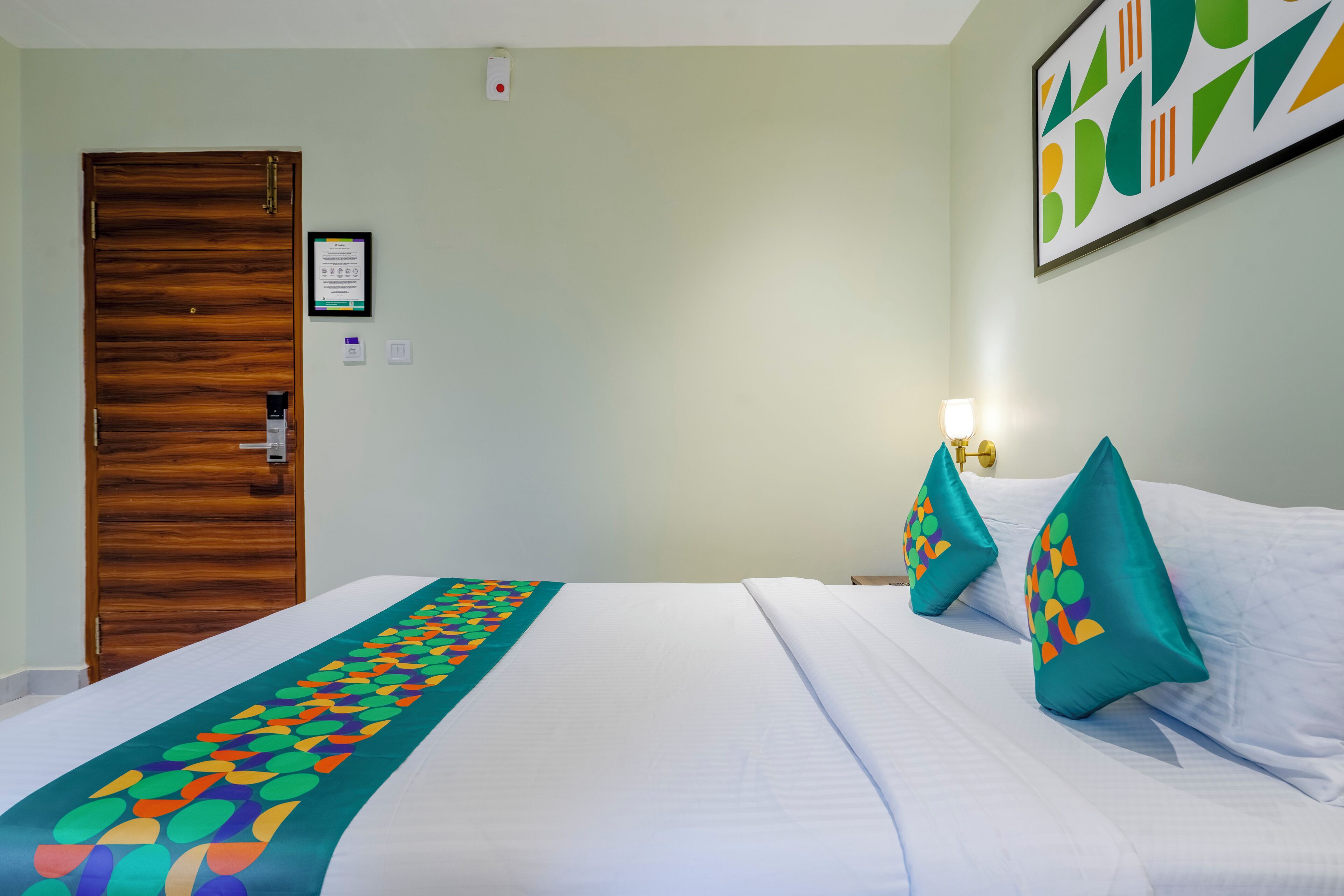 A hotel room with a wooden door, a bed with white bedding and colorful pillows, a framed certificate on the wall, and a wall-mounted light.