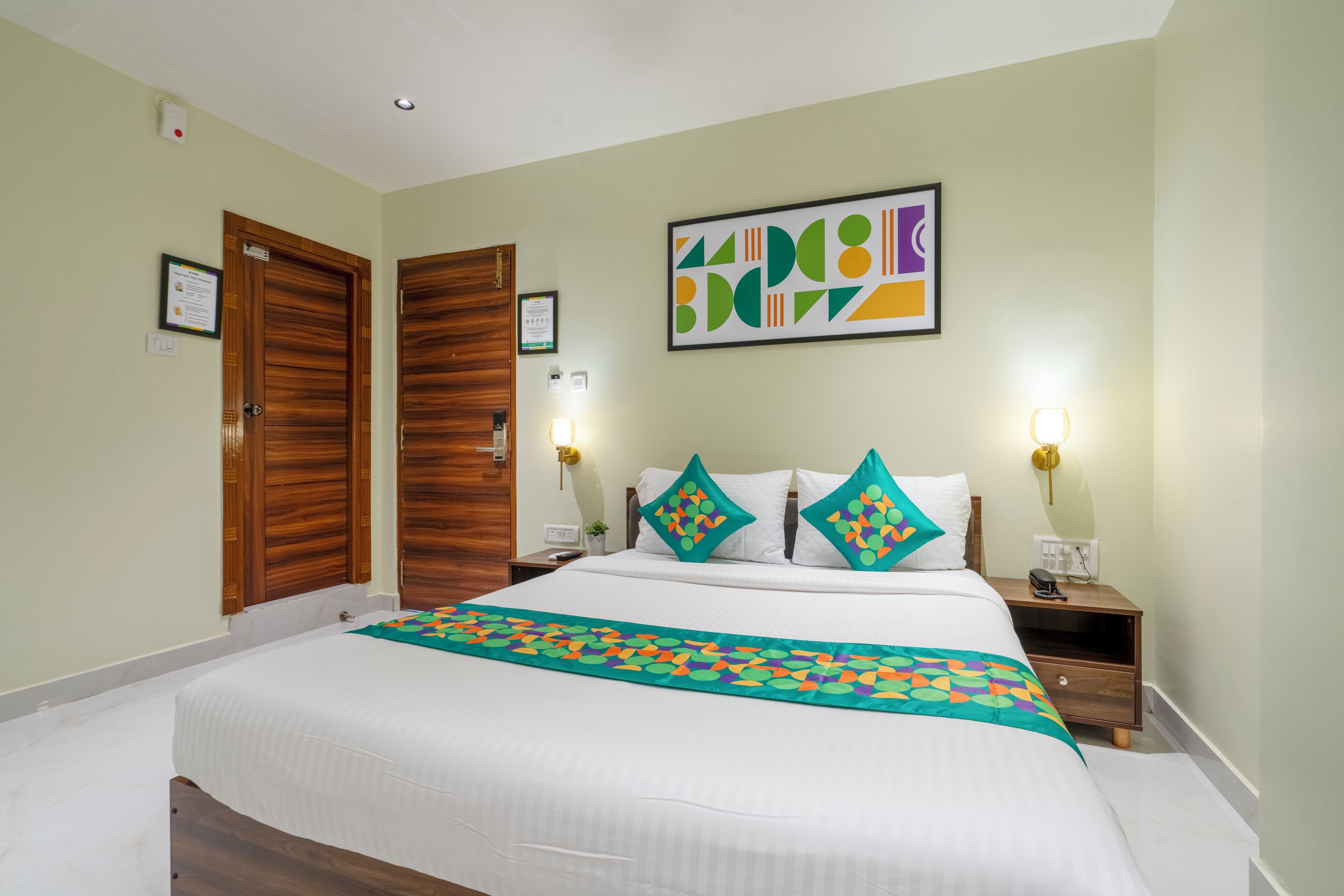 Habitación de hotel con una cama grande, dos puertas de madera, un cuadro enmarcado en la pared y una mesita de noche con una lámpara.