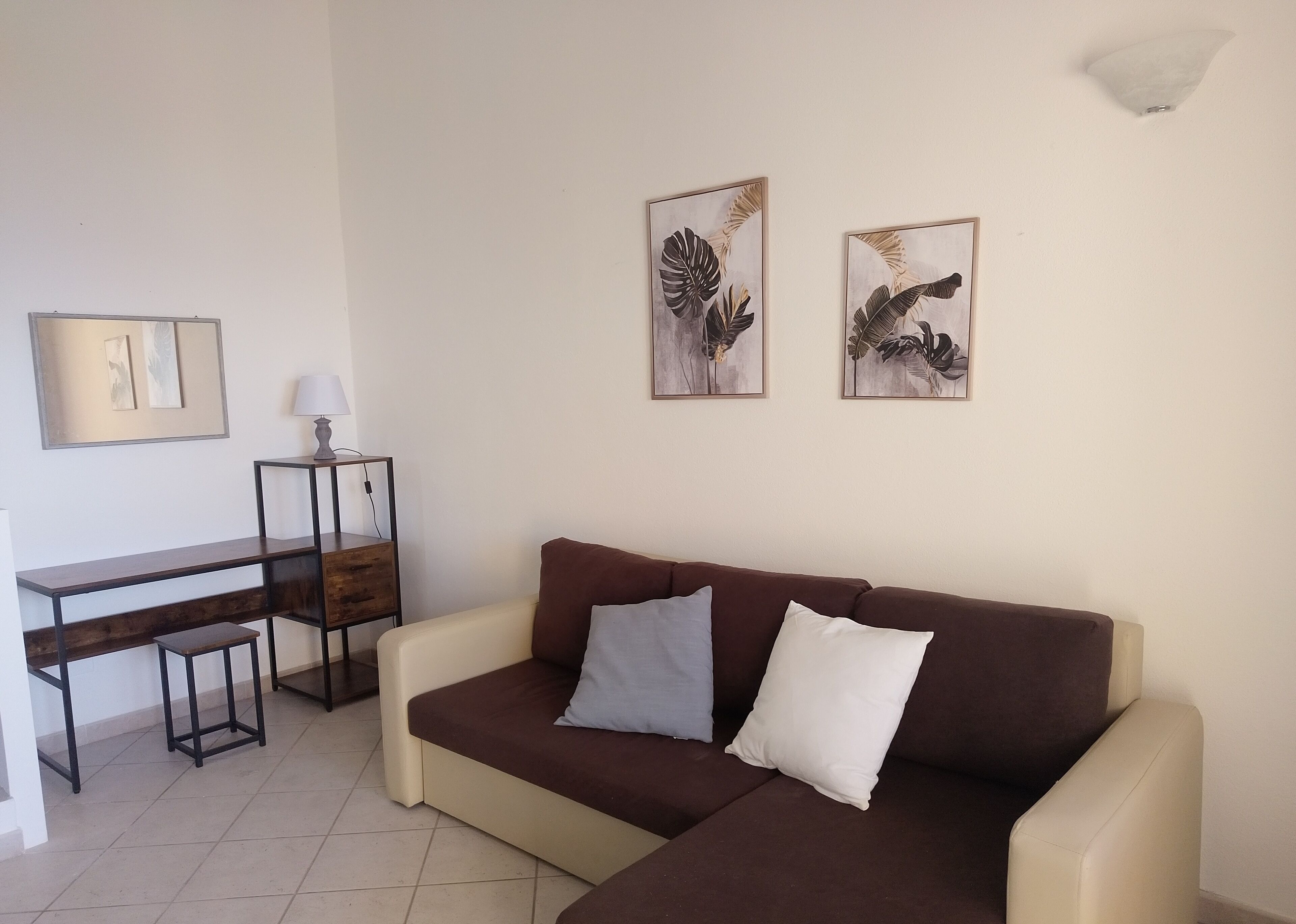 Un salon avec un canapé marron, deux coussins, une petite table avec une lampe et deux œuvres d’art représentant des feuilles, encadrées et accrochées au mur.