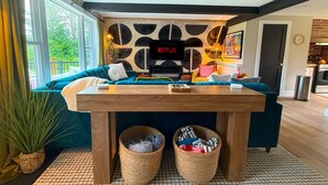 Una sala de estar moderna con un sofá grande, una mesa de centro de madera y un televisor empotrado que muestra 'NETFLIX'. Hay dos cestas debajo de la mesa y una planta en maceta a la izquierda.