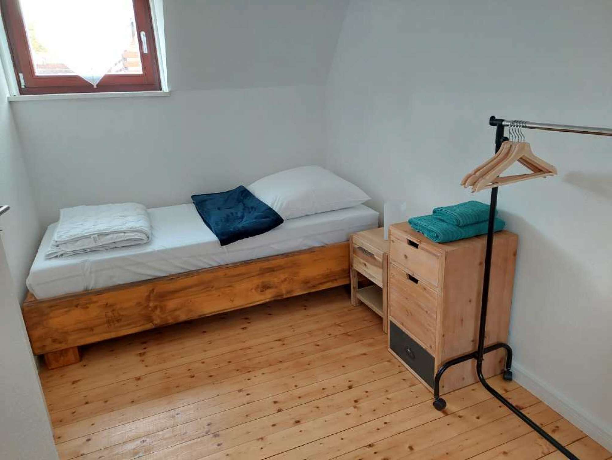 Une petite chambre avec un lit simple, un plancher en bois, un lit blanc, une table de chevet en bois, un portant à vêtements et une fenêtre donnant sur un paysage agréable.