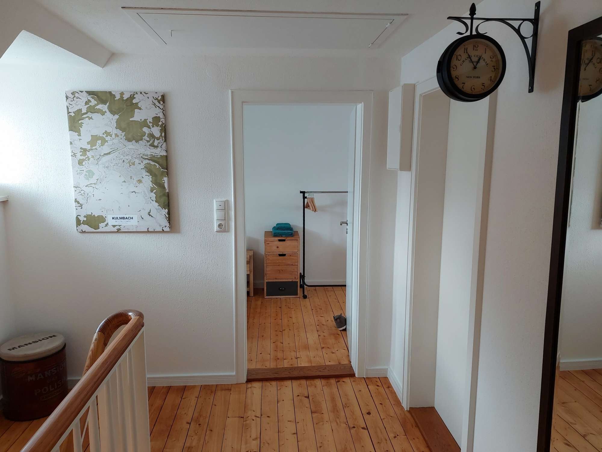 Un pasillo con suelo de madera, un mapa en la pared, un reloj y una puerta que da a otra habitación.