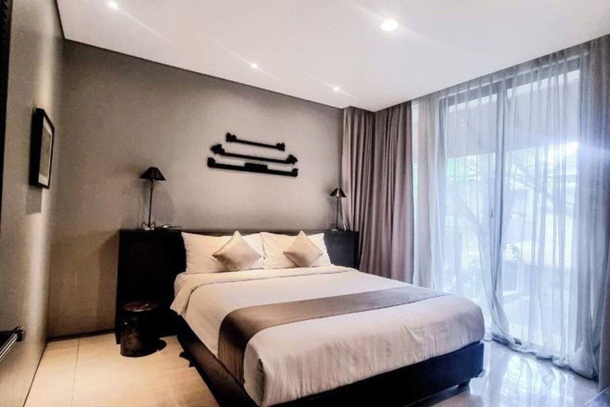 A modern bedroom with a large bed, two bedside tables, and a view of the outdoors through sheer curtains.