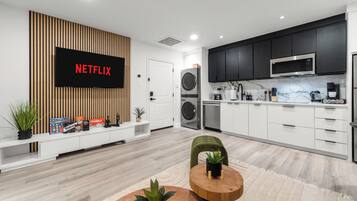 A modern living room with a flat-screen TV, a wooden wall panel, a white entertainment unit, and a round wooden coffee table with a plant.