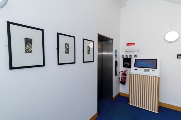 A hallway with framed artwork on the wall, a fire extinguisher, a fire alarm, and a digital information kiosk.