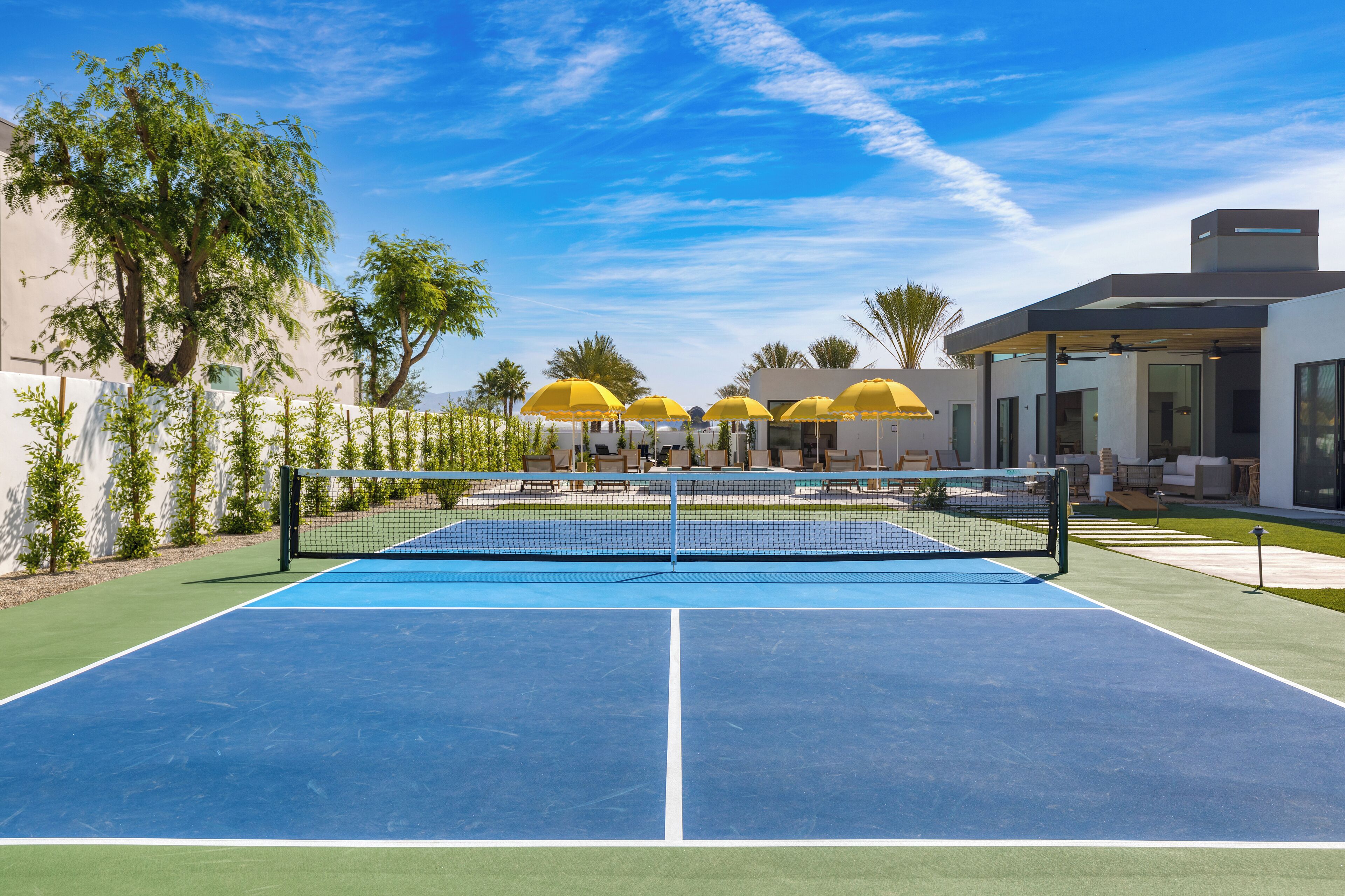 Una cancha de tenis exterior con red, rodeada de palmeras y sombrillas amarillas.