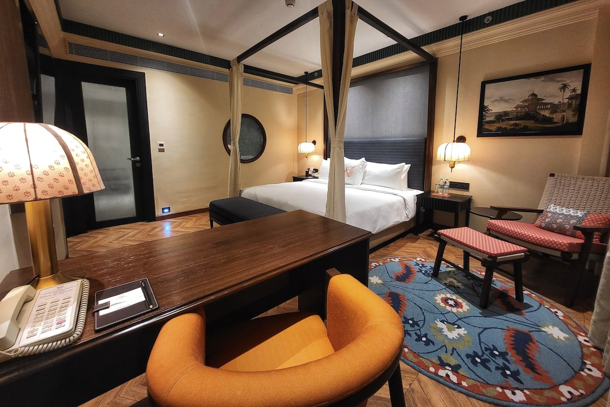 Habitación de hotel con cama, escritorio con teléfono, silla, mesita, mesita de noche, lámpara, alfombra y un cuadro en la pared.