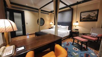 Habitación de hotel con cama, escritorio con teléfono, silla, mesita, mesita de noche, lámpara, alfombra y un cuadro en la pared.