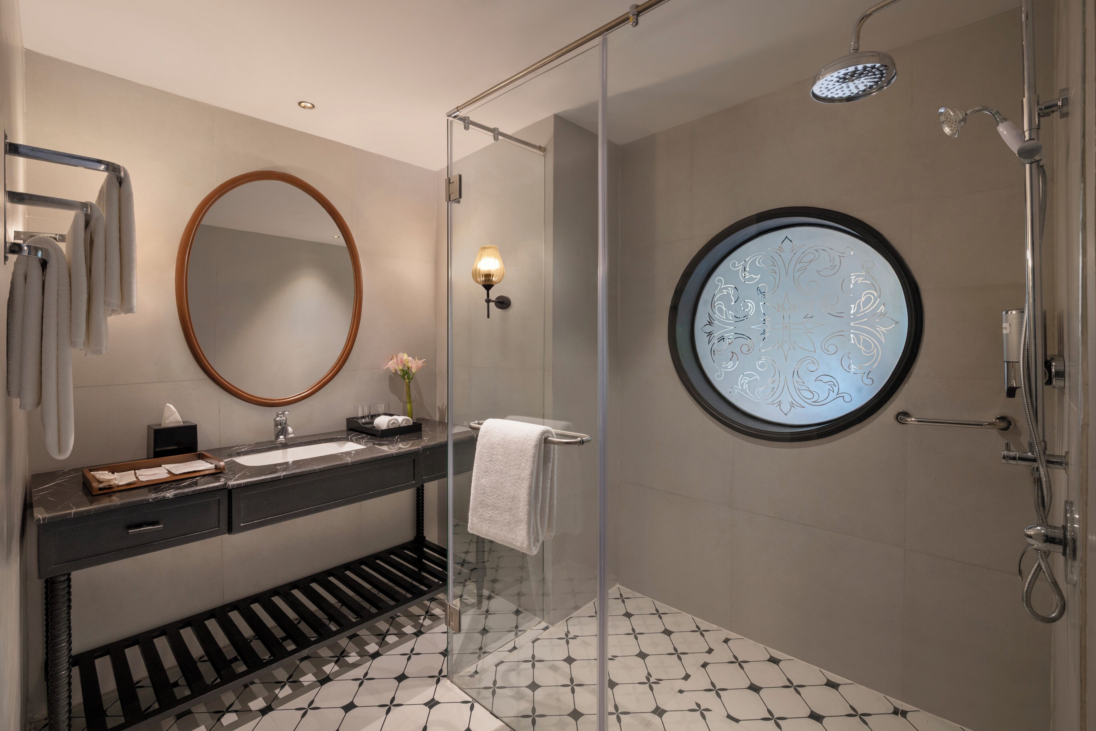 Un baño moderno con cabina de ducha de vidrio, un espejo redondo, tocador con encimera de mármol y suelo estampado.