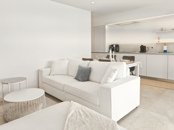 A modern living room with a white sofa, a round side table, and a dining area in the background. - Modern Pool Bungalow (Torrevieja)