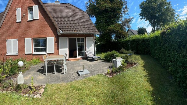 A red brick house with a tiled roof, a patio with a table and chairs, and a garden with a hedge and trees. - Strandgut - Half of a house in the center of Zingst (Zingst)