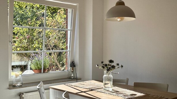 A dining area with a table set for two, a hanging light fixture, and a window with a view of greenery. - Strandgut - Half of a house in the center of Zingst (Zingst)