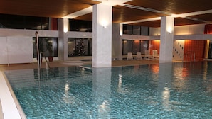 Una piscina interior con agua cristalina, rodeada de paredes y columnas blancas, y un área de descanso con sillas.