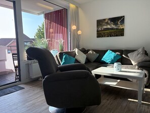 Una sala de estar moderna con sofá, sillón y mesa de centro. Hay una ventana con cortinas, un cuadro en la pared y un radiador.