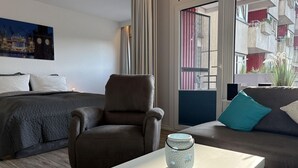 Una habitación de hotel moderna con una cama, un sillón marrón, una mesa blanca y un sofá con cojines verde azulado y blancos.