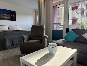 Una habitación de hotel moderna con una cama, un sillón marrón, una mesa blanca y un sofá con cojines verde azulado y blancos.