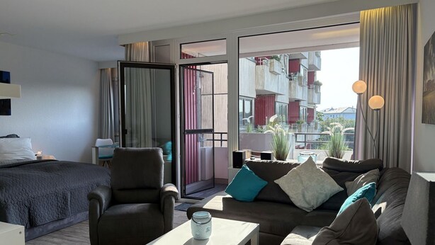 Una habitación de hotel moderna con cama, sofá, silla, una mesita y balcón con vistas a la ciudad.
