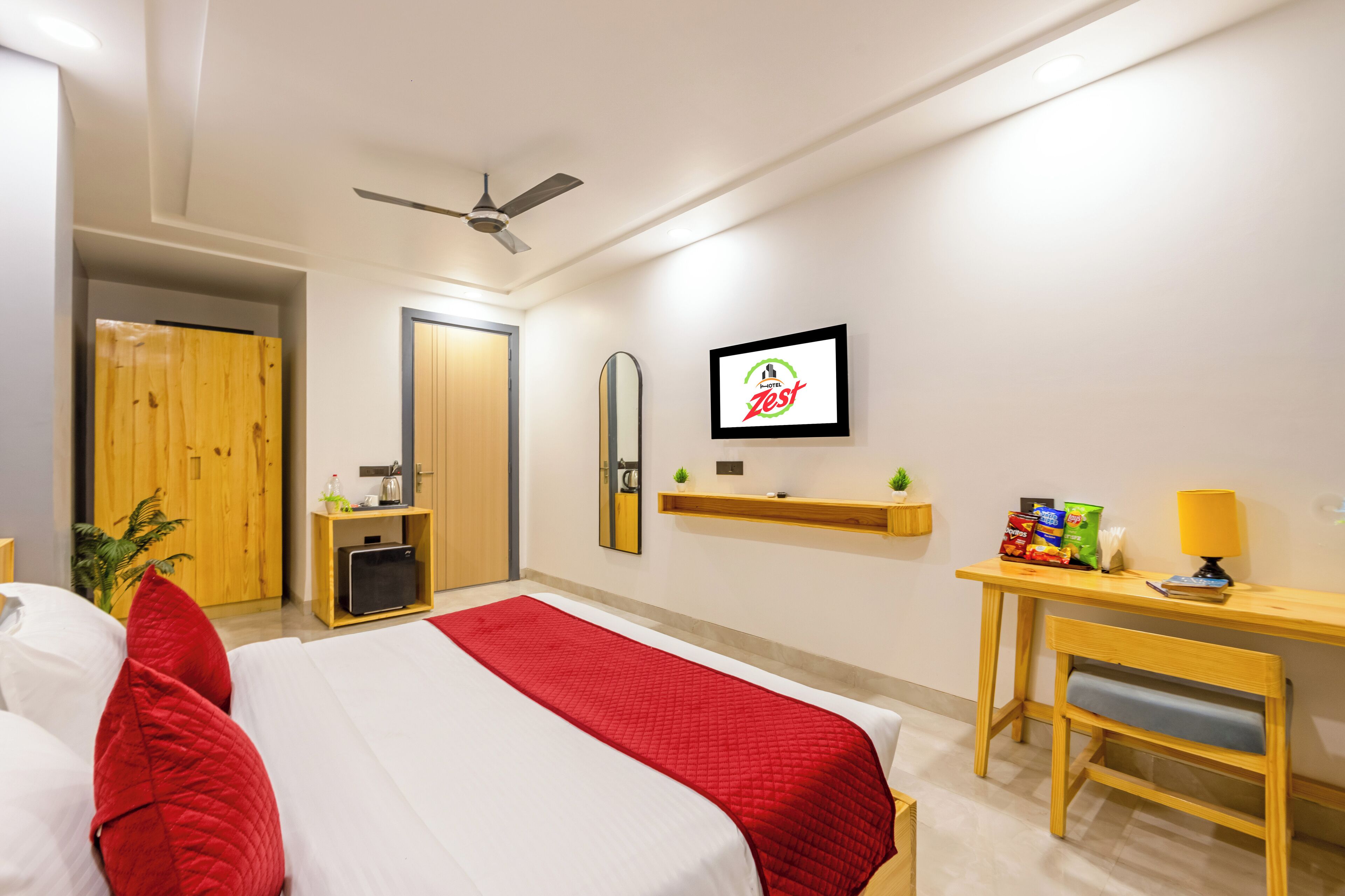 A hotel room with a bed, a desk, a chair, a ceiling fan, a mirror, and a shelf with items.