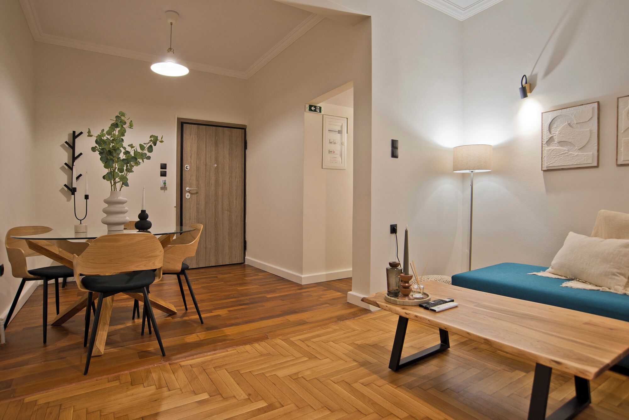 Uma sala de estar moderna com piso de madeira, um sofá azul, uma mesa de centro de madeira e uma área de jantar com mesa de vidro e cadeiras de madeira.