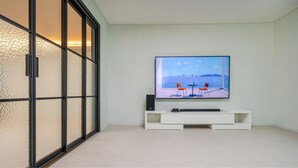 Una sala moderna con un televisor de pantalla plana grande montado en la pared, un mueble blanco de entretenimiento debajo y un ventanal corredero que da a un balcón con vistas al mar.