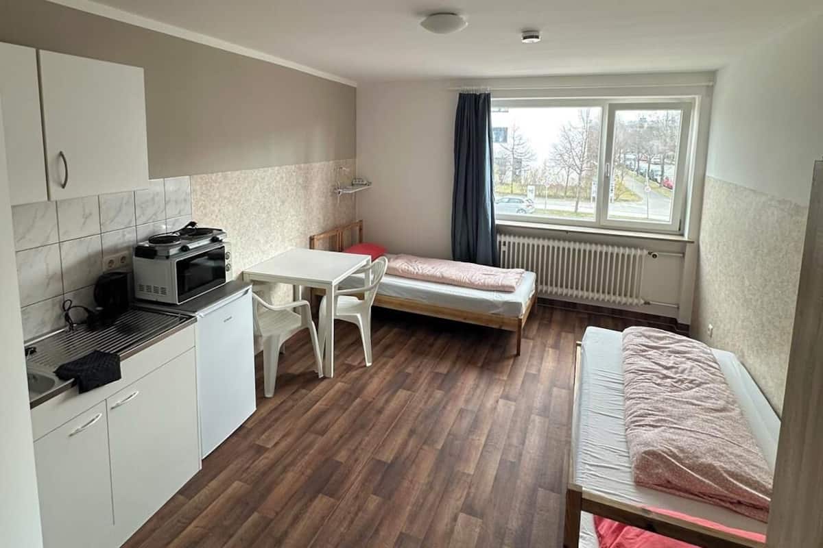 Una habitación pequeña con kitchenette, una cama individual, una mesita y una ventana.