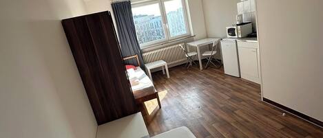 A compact room with a bed, a small table, a kitchenette, and a view of the city through a window.