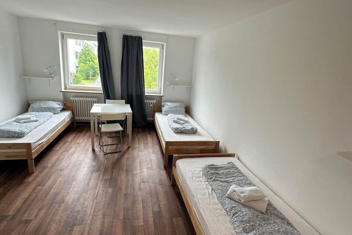 Una habitación tipo dormitorio con dos camas individuales, una mesita y un ventanal con cortinas.