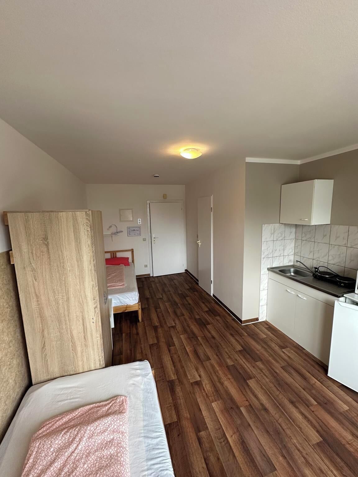 A compact room with a bed, a kitchenette, and a door leading to another room.