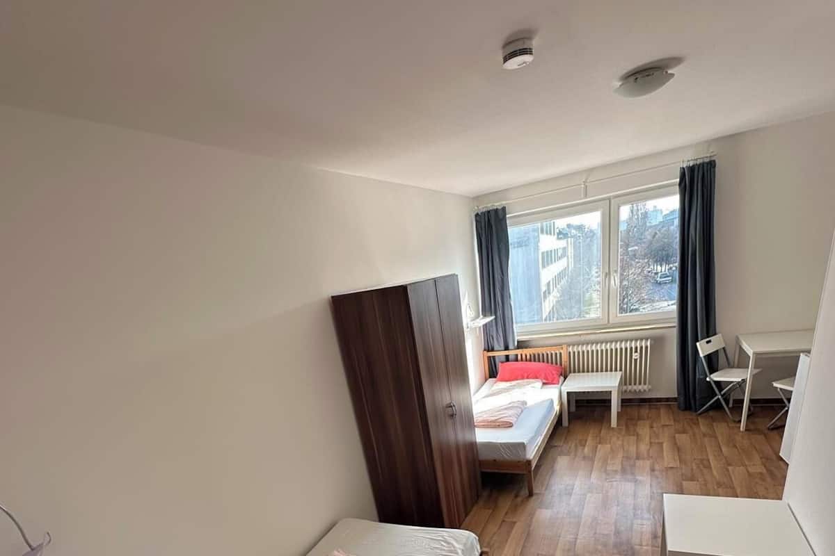 Un dormitorio con dos camas individuales, suelo de madera, ventana con cortinas, un radiador y un escritorio blanco.