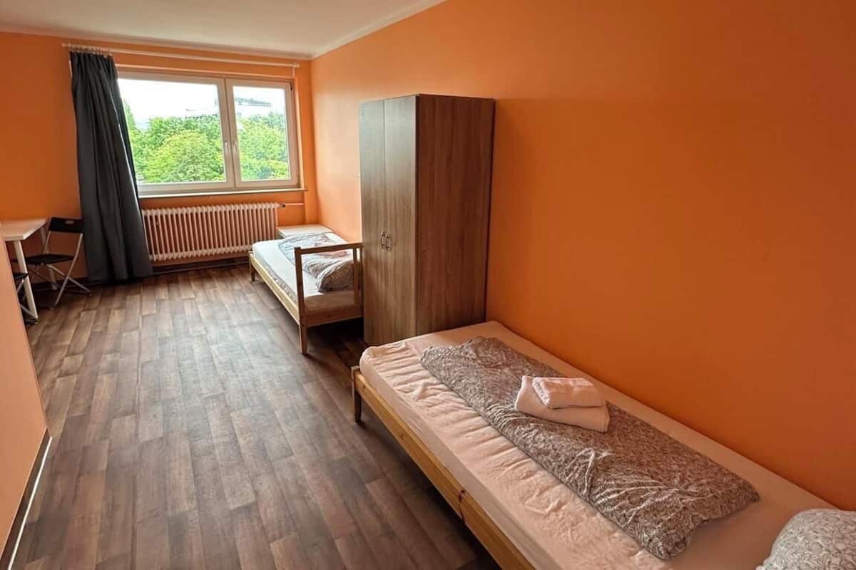 Habitación individual con piso de madera, una ventana con cortinas, un radiador y un armario.