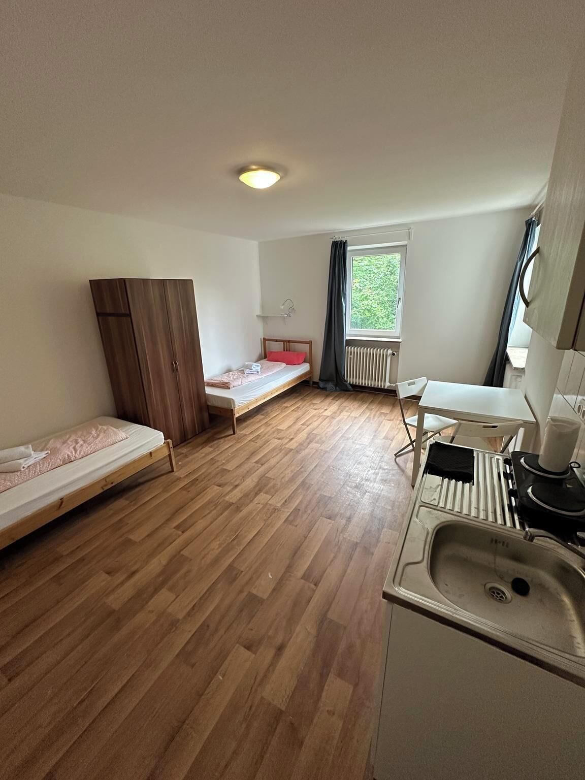 Habitación pequeña con dos camas individuales, un armario de madera, una mesita, un lavamanos y una ventana con vistas a zonas verdes.