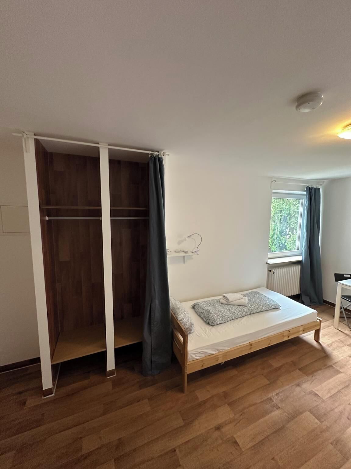 Una habitación pequeña con cama individual, suelo de madera, ventana con cortinas, escritorio y armario con puertas correderas.