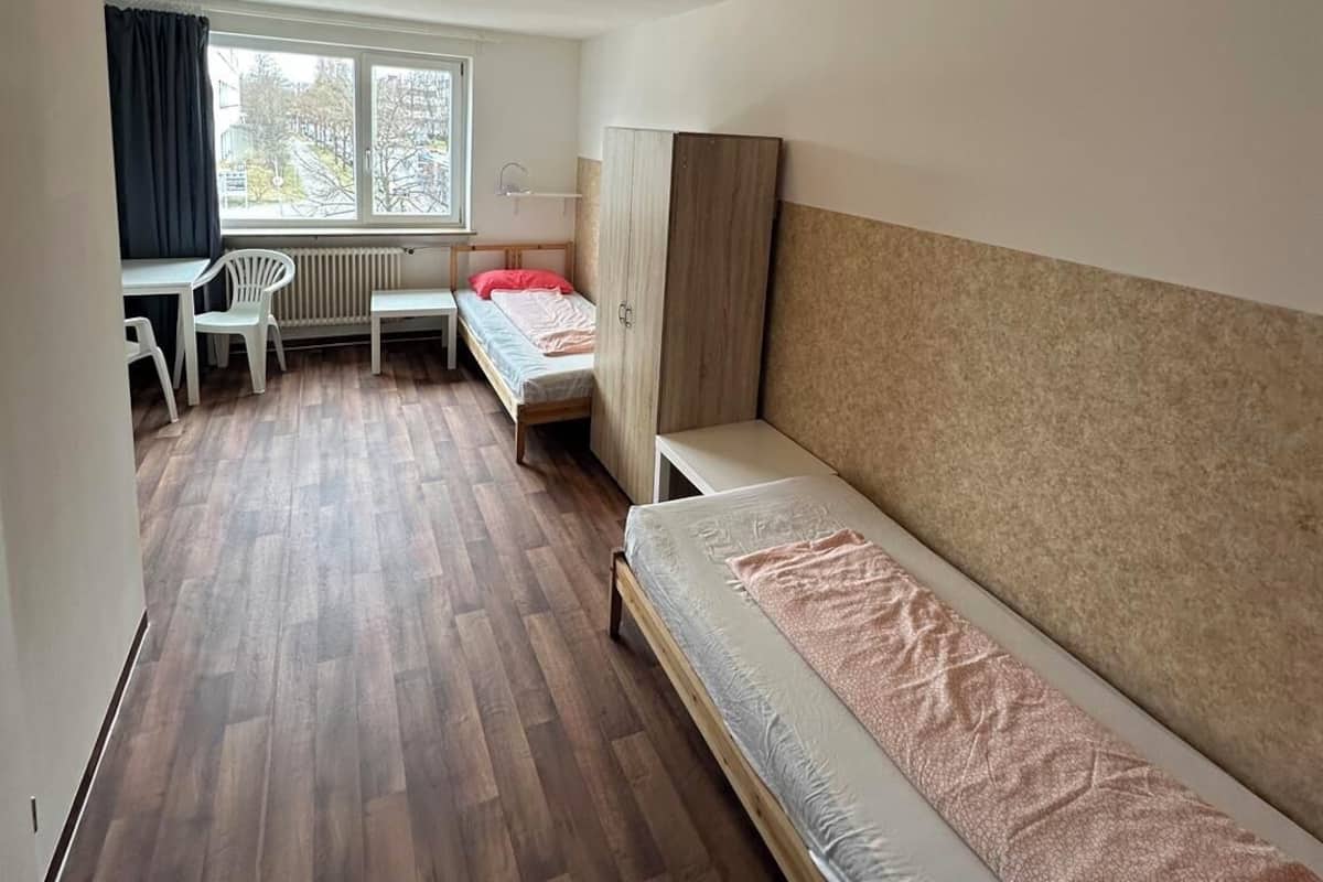 Una habitación pequeña y bien iluminada, con piso de madera, una cama individual, un escritorio con silla y una ventana con cortinas.