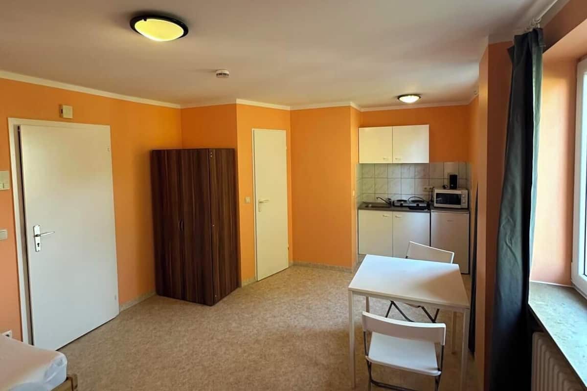 Una habitación con dos camas, una mesita, una silla, un rincón americano y una ventana con cortinas.