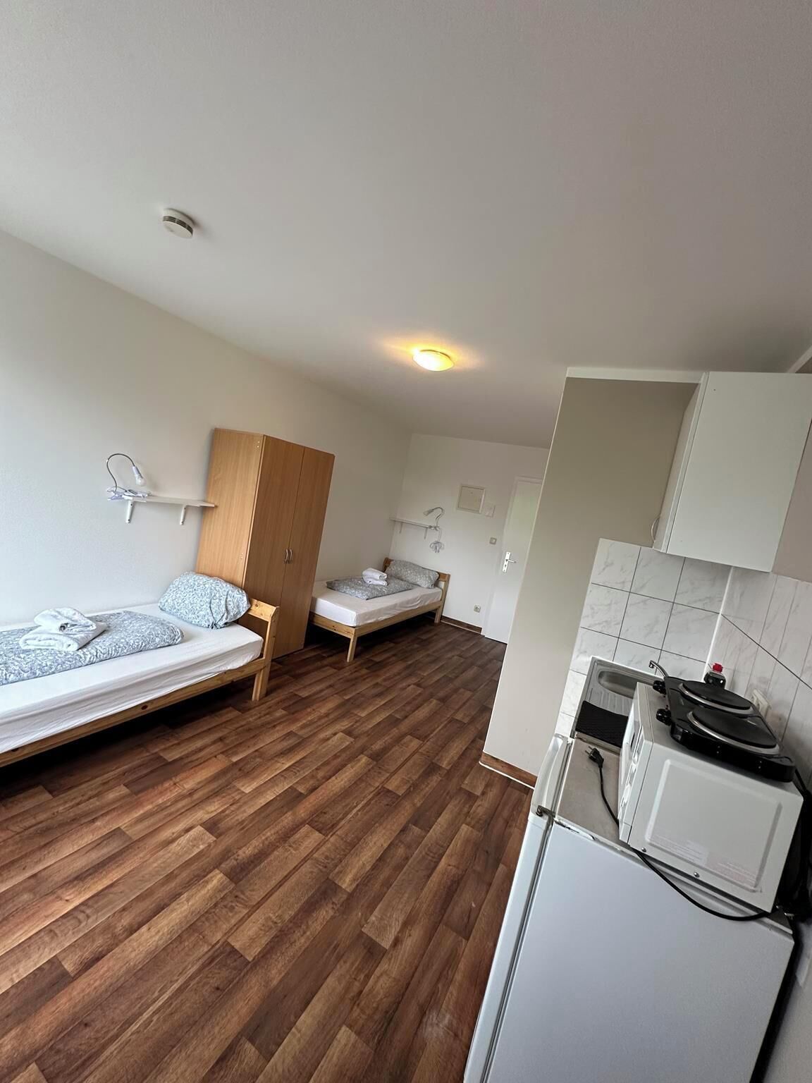 A compact room with two single beds, a wooden wardrobe, a small kitchenette with a fridge and stove, and a tiled floor.