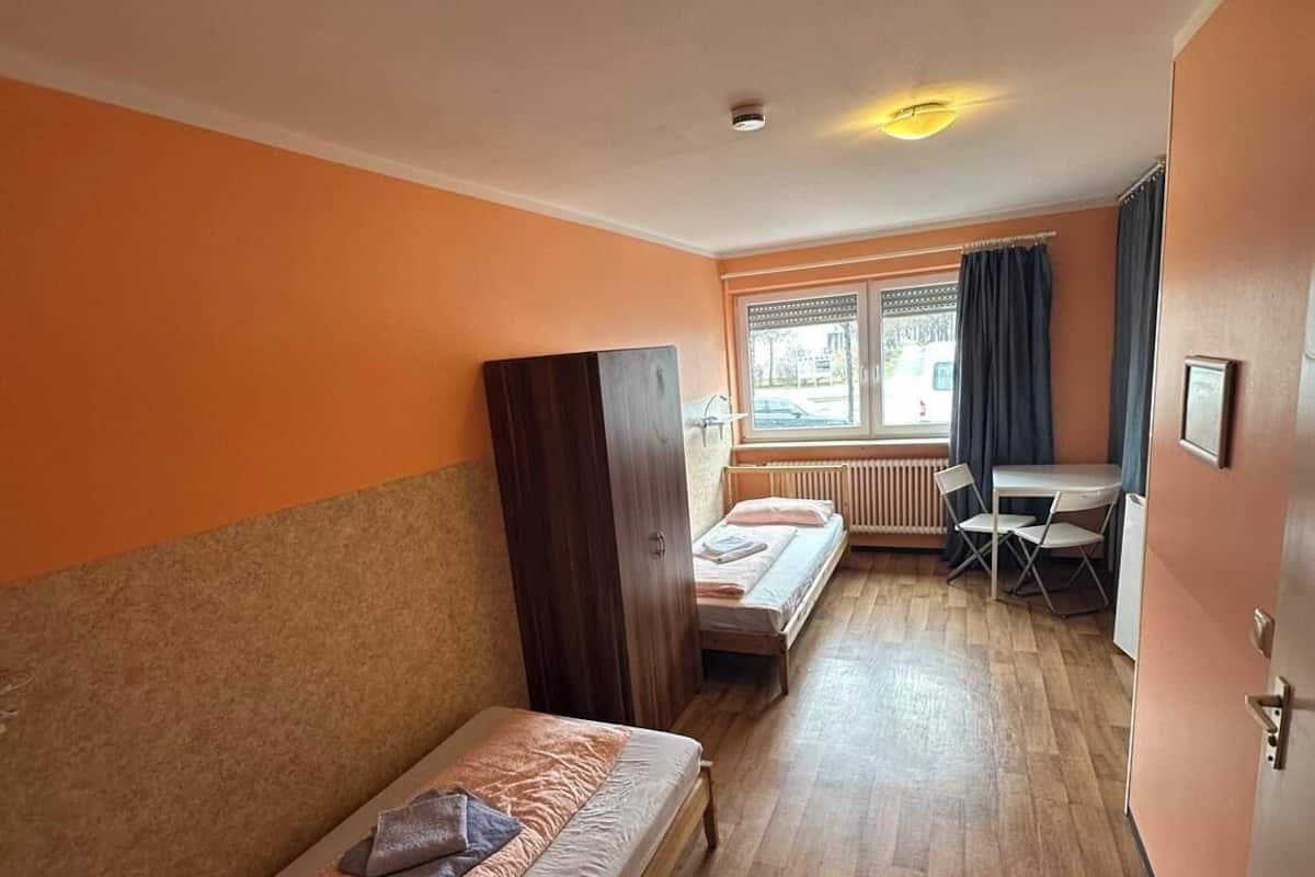 Una habitación con dos camas individuales, un escritorio, una silla, un radiador y una ventana con cortinas.