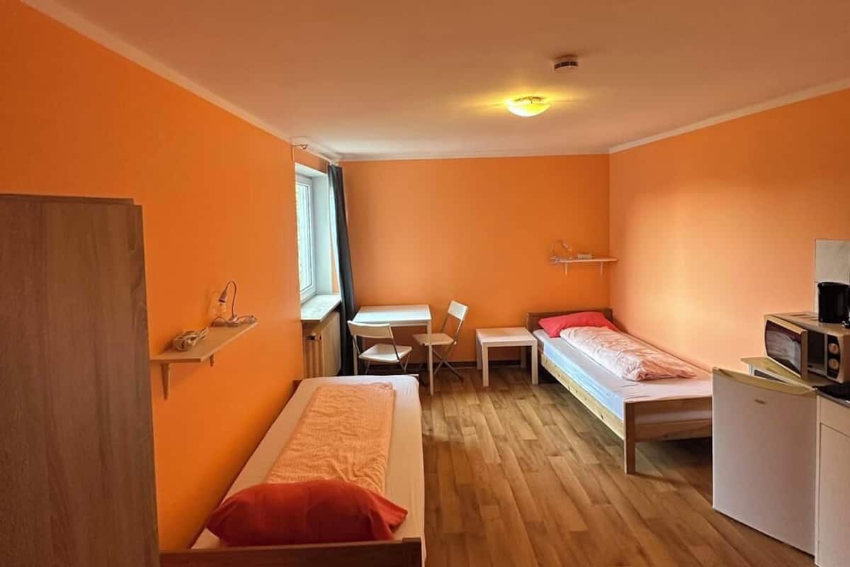 Una habitación pequeña con dos camas individuales, una mesita, una silla, un escritorio y un microondas.