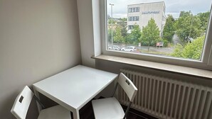 Une petite pièce avec une table blanche et deux chaises, un radiateur et une fenêtre donnant sur un immeuble et des arbres.
